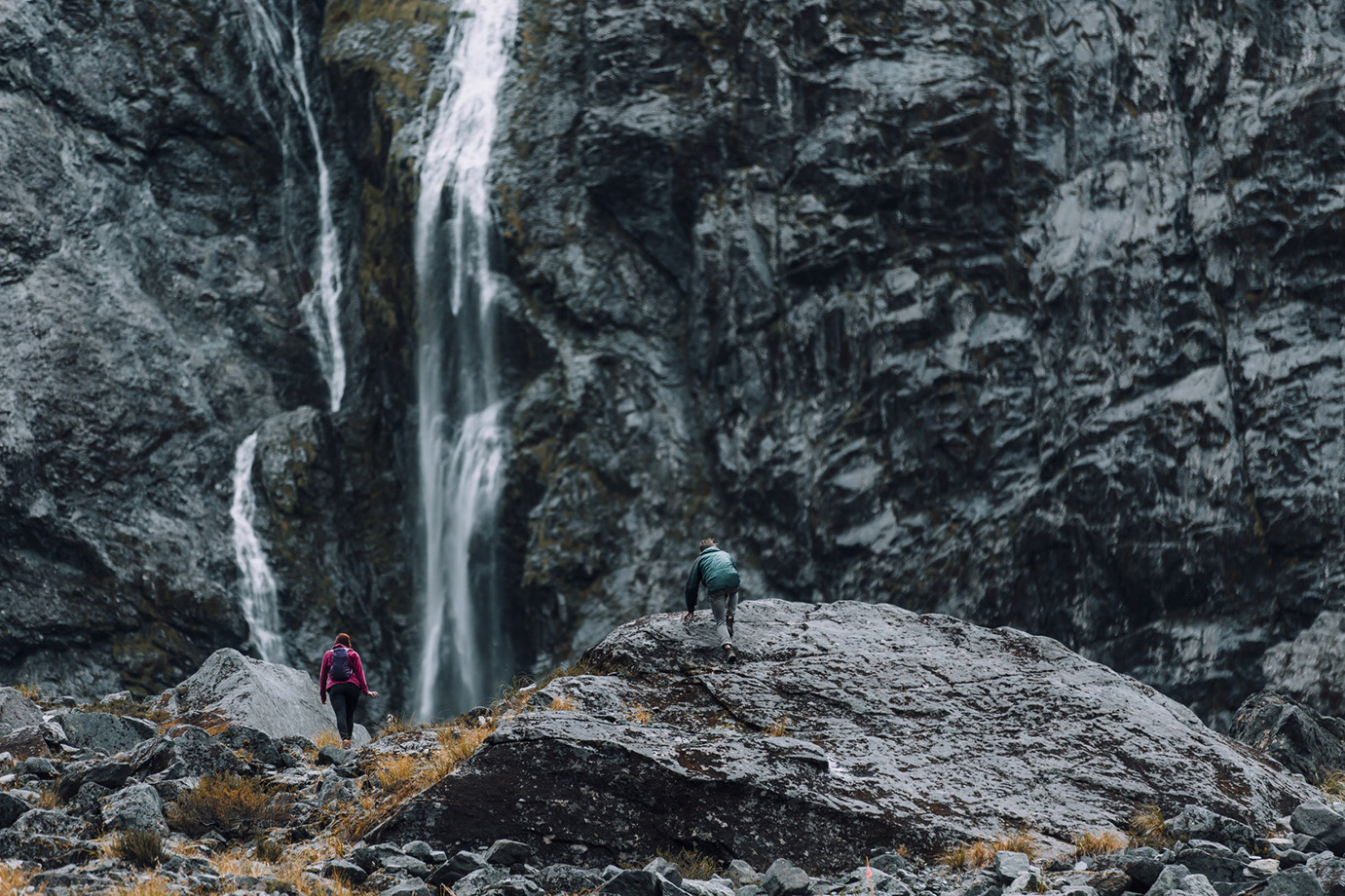 An athletic couple climbs rocky terrain to the base of a waterfall in New Zealand