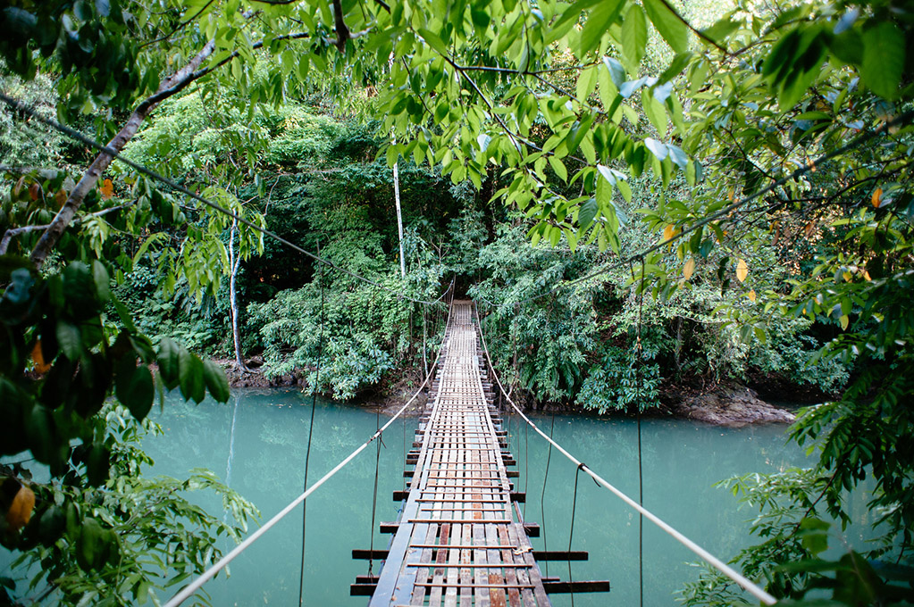 A suspension bridge crossing a river through dense tropical jungle — the road to Pavones has several of these, along with the potholes that make every hour after the last sealed section feel earned