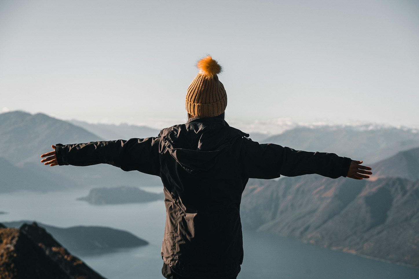 A figure on a mountain summit at golden hour, arms open to the valley below — the view that makes every kilometre of approach and every gram of camera kit worth carrying